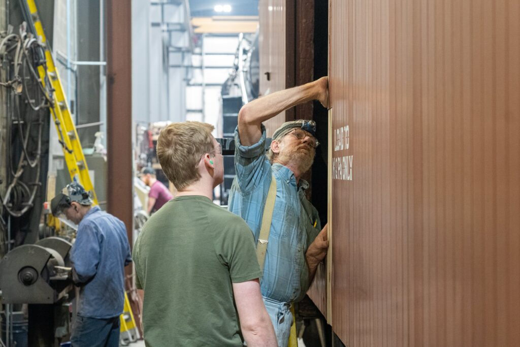 Two men cosmetically restoring a steam locomotive in a workshop on the door of a railcar